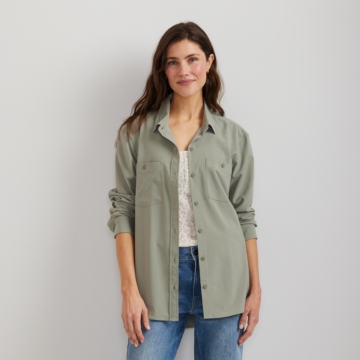 Sage green long-sleeve button-up shirt with chest pockets, worn open over a patterned white top and blue jeans.
