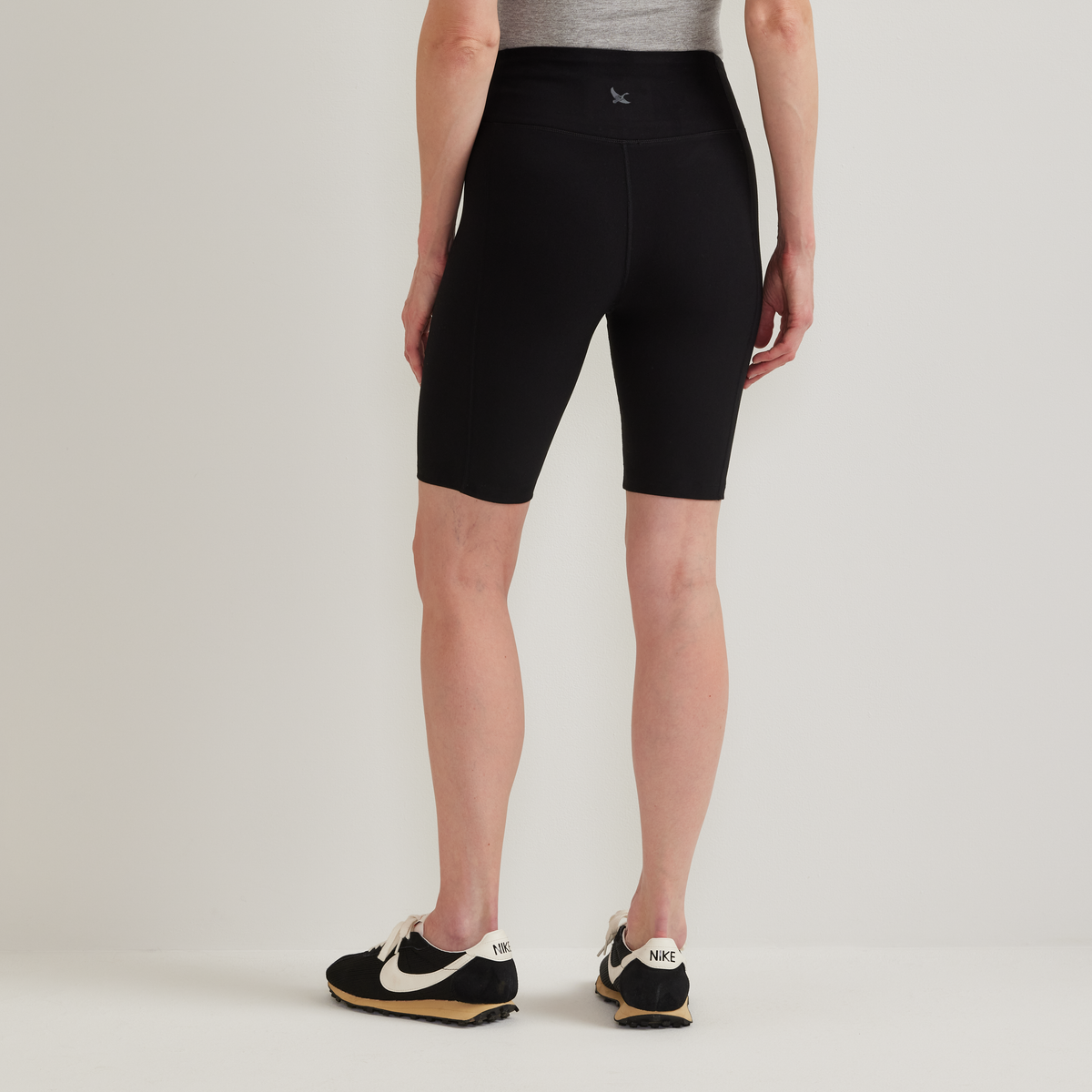 Black knee-length bike shorts with high waistband on woman wearing gray top and black sneakers.
