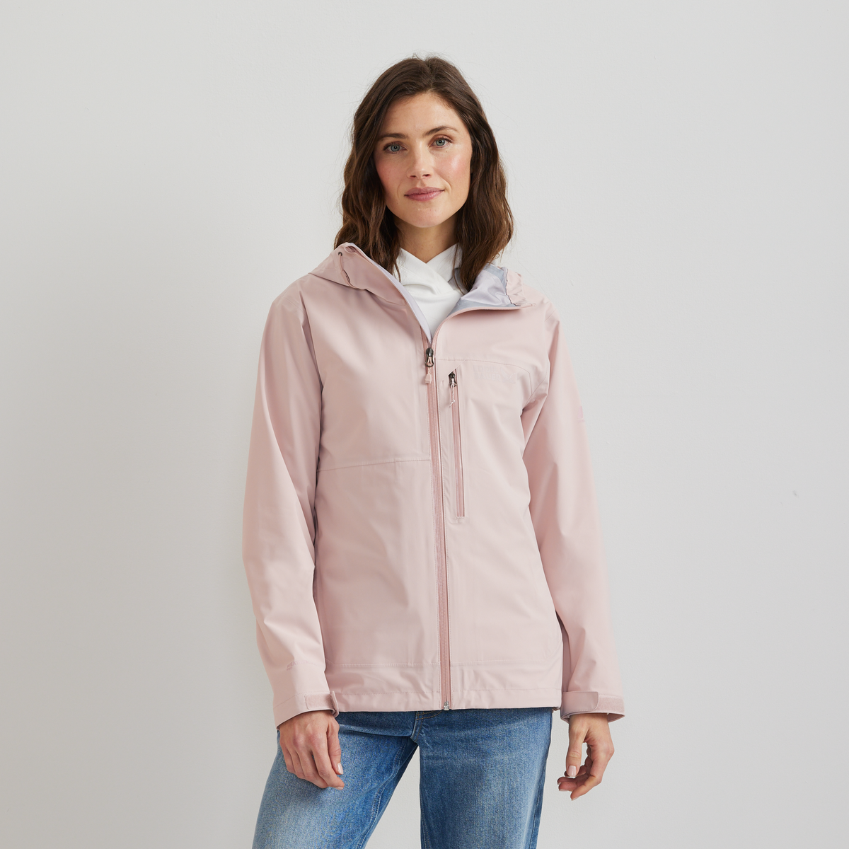 Woman wearing a light pink hooded rain jacket with front zipper over a white collared shirt and blue jeans.