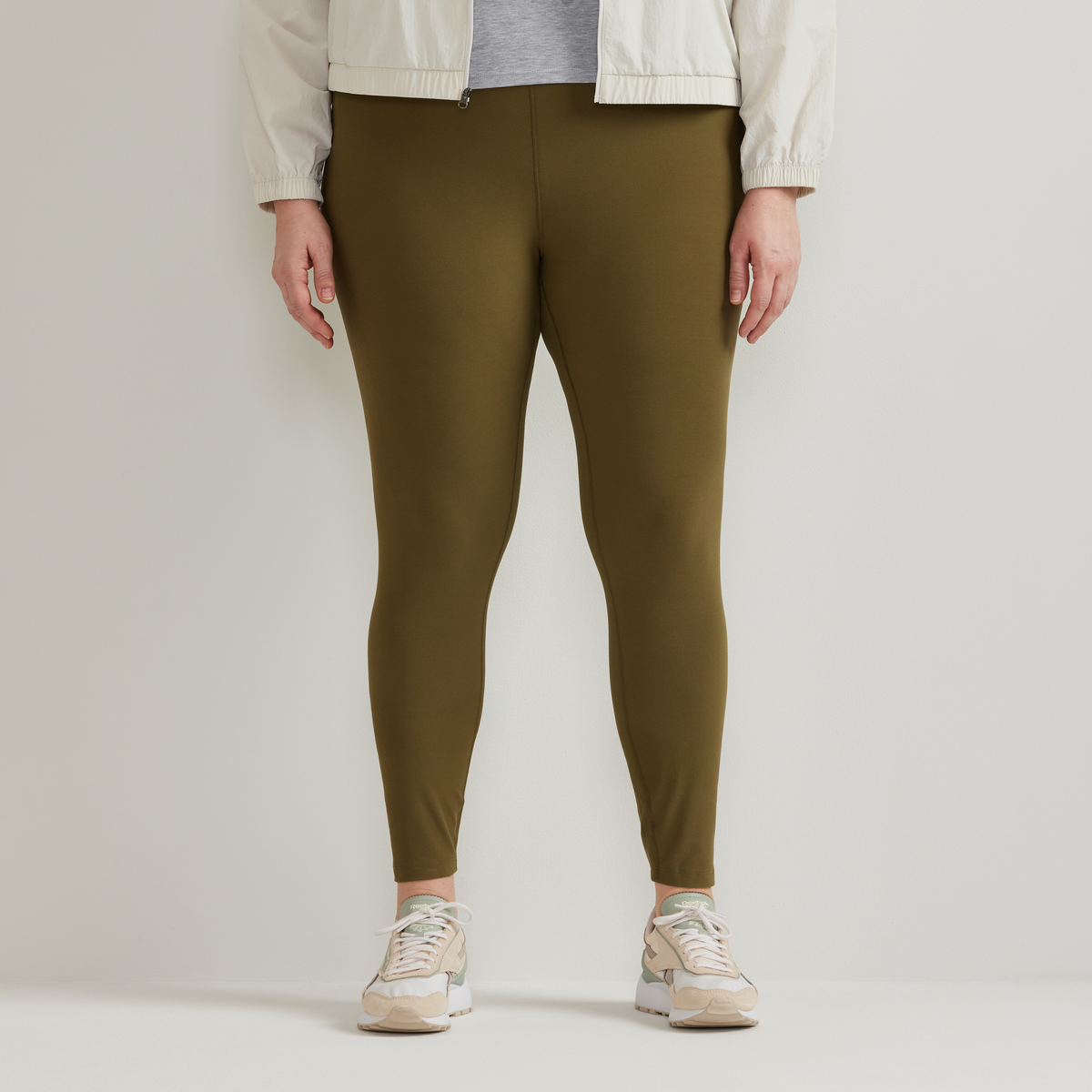 Olive green high-rise leggings with fitted silhouette, shown on model wearing white jacket and beige sneakers.