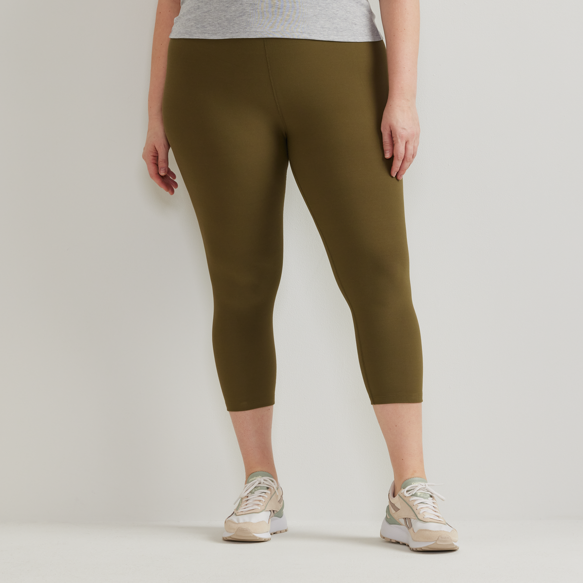Olive green high-rise capri leggings on a model wearing a gray tank top and beige sneakers.